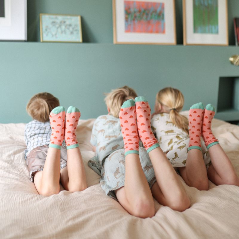Chaussettes sans couture à motifs cœurs rouge de différentes tailles portées par trois enfants qui posent sur un lit.