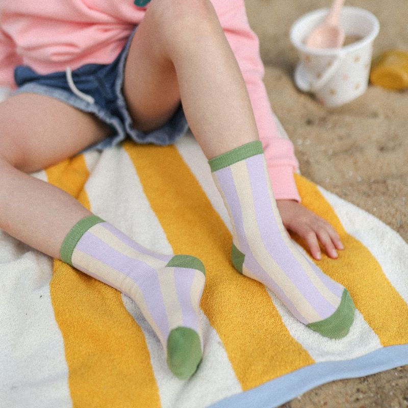 Chaussettes sans couture rayées vert et mauve portées par un enfant avec une mise en ambiance plage