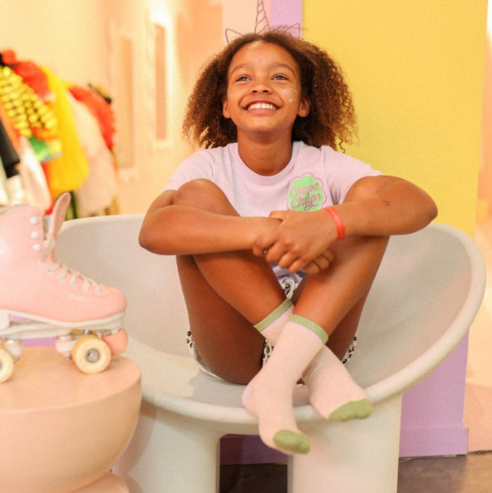 Photo des chaussettes sans couture unies paillettes rose pale et vert portées par une enfant assise dans une ambiance colorée avec des patins à roulettes.