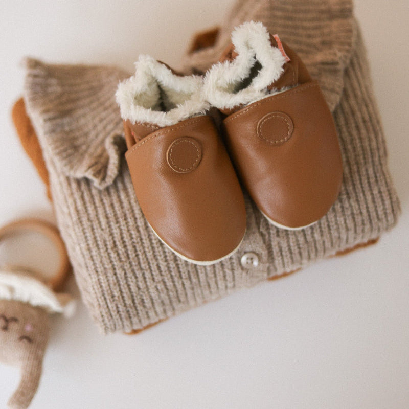 Les chaussons cuir souple fourrés bébé dans une mise en ambiance de chambre de bébé.