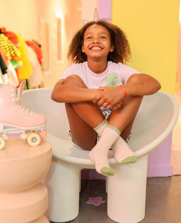 Photo des chaussettes sans couture unies paillettes rose pale et vert portées par une enfant assise dans une ambiance colorée avec des patins à roulettes.