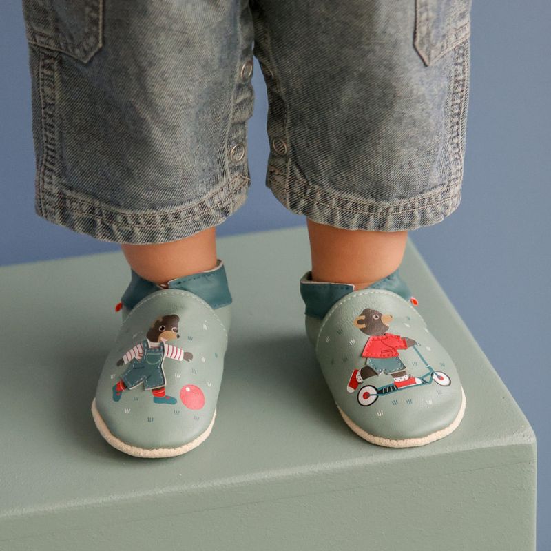 Chaussons bleu en cuir souple avec des motifs Petit Ours Brun qui joue et portés par une bébé. 